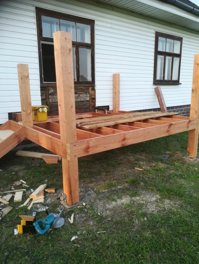 Drewniana konstrukcja tarasu w trakcie budowy przy białym domu z oknami, widoczne narzędzia i resztki drewna na trawie.