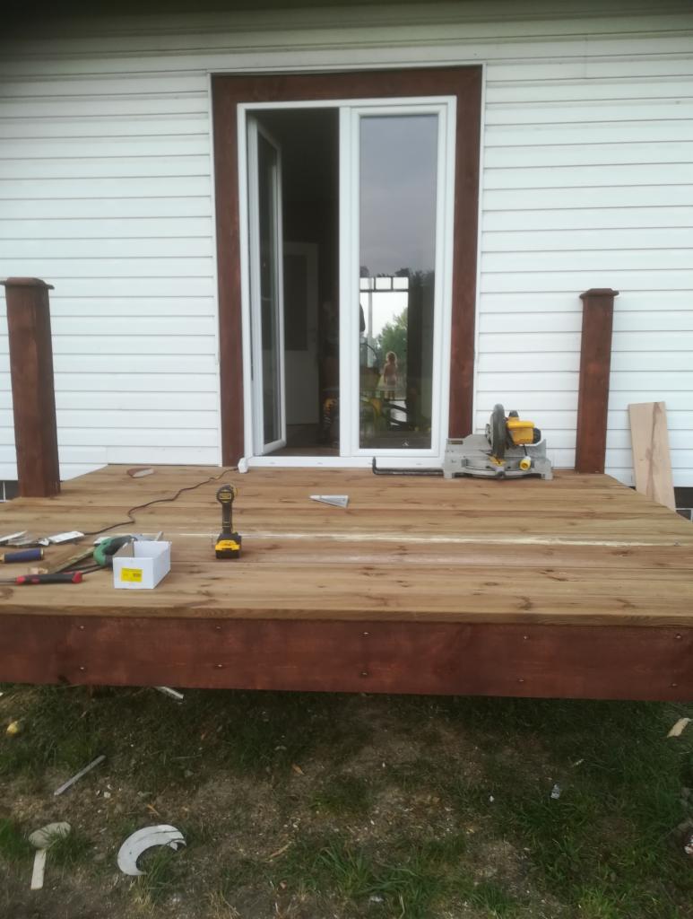 Budowa drewnianego tarasu przed domem z białym sidingiem, widoczna otwarta, przeszklona rama drzwiowa z brązowym obramowaniem, narzędzia pracy: wiertarka, piła, poziomica.