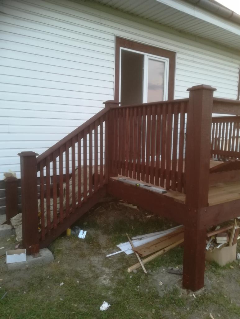 Brązowe schody i taras przed domem z białym sidingiem, widoczne narzędzia i materiały budowlane na ziemi, częściowo ukończona konstrukcja.