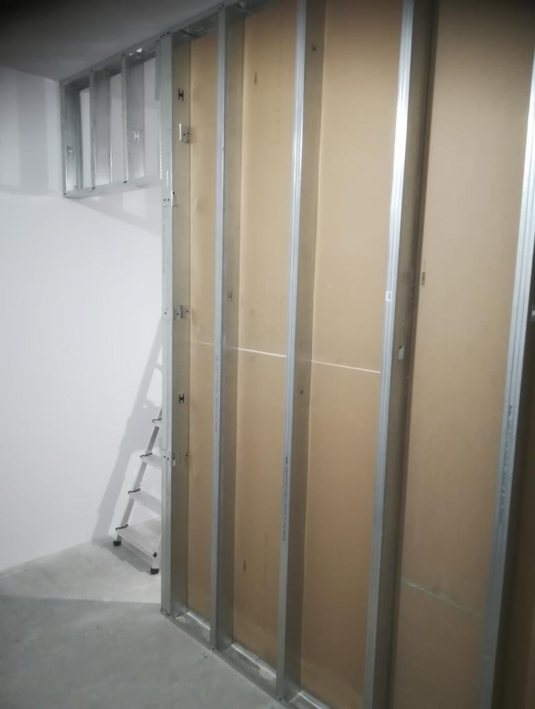 Konstrukcja ściany działowej z profili metalowych i płyt kartonowo-gipsowych w trakcie montażu; widoczna drabina oparta o ścianę w surowym stanie.