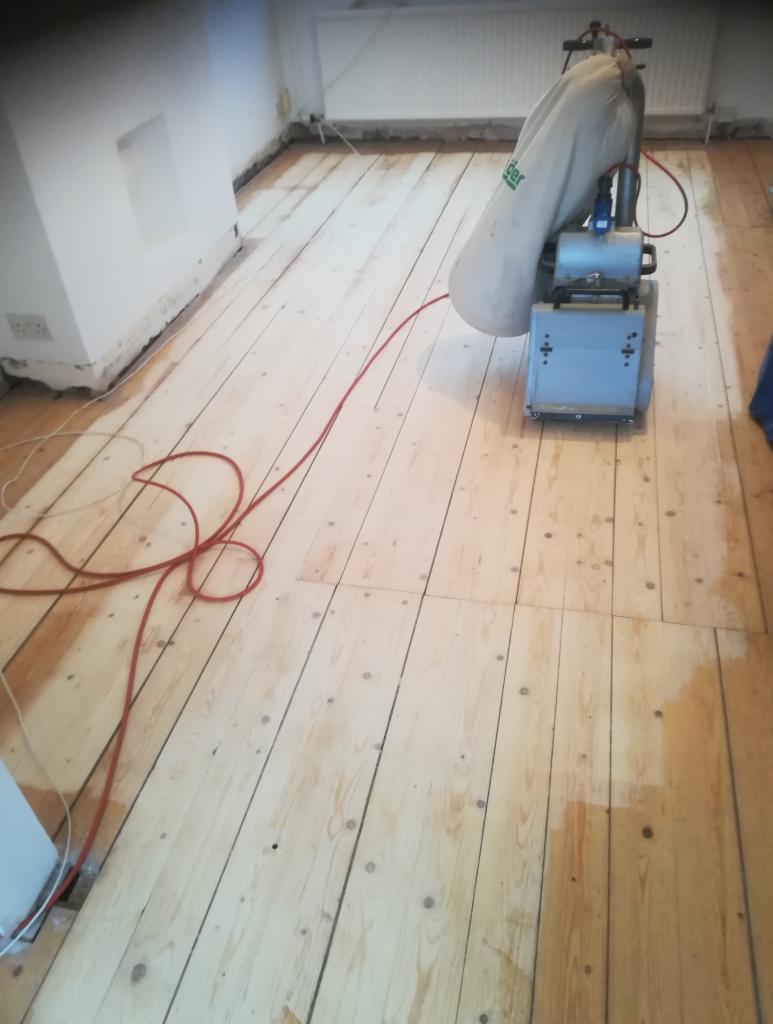 Parkiet drewniany w trakcie cyklinowania, widoczna maszyna do cyklinowania i fragment wycyklinowanej powierzchni podłogi.