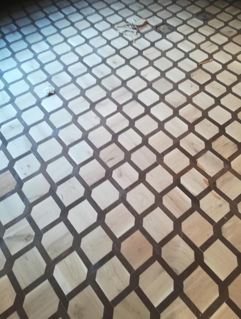Parkiet ułożony w geometryczny wzór z jasnego drewna i ciemnych wstawek, widoczne drobne zabrudzenia i pozostałości po pracy.