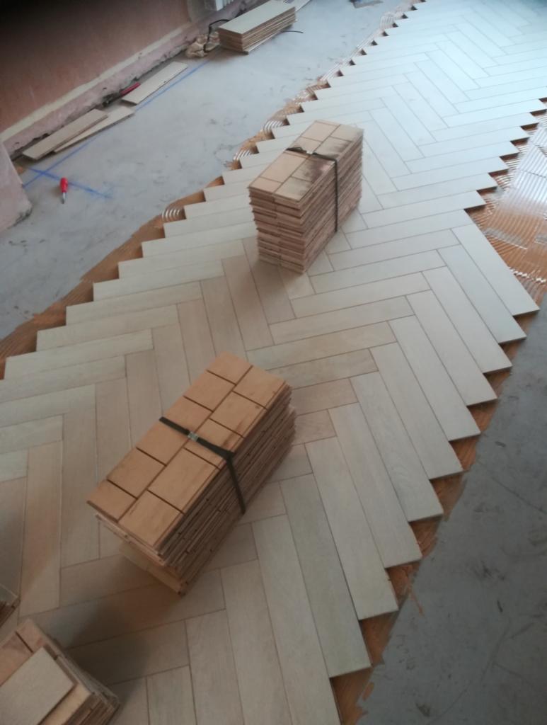 Układanie jasnych paneli podłogowych w jodełkę, widoczne stosy paneli gotowych do montażu i fragment podłoża z klejem.