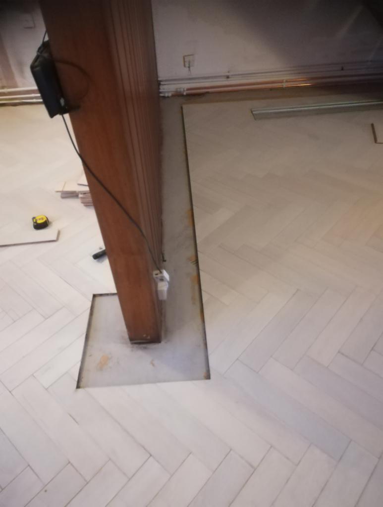 Układanie parkietu w jodełkę wokół drewnianej kolumny, z widocznym wycięciem w podłodze wokół podstawy kolumny, w tle rury instalacyjne pod sufitem.