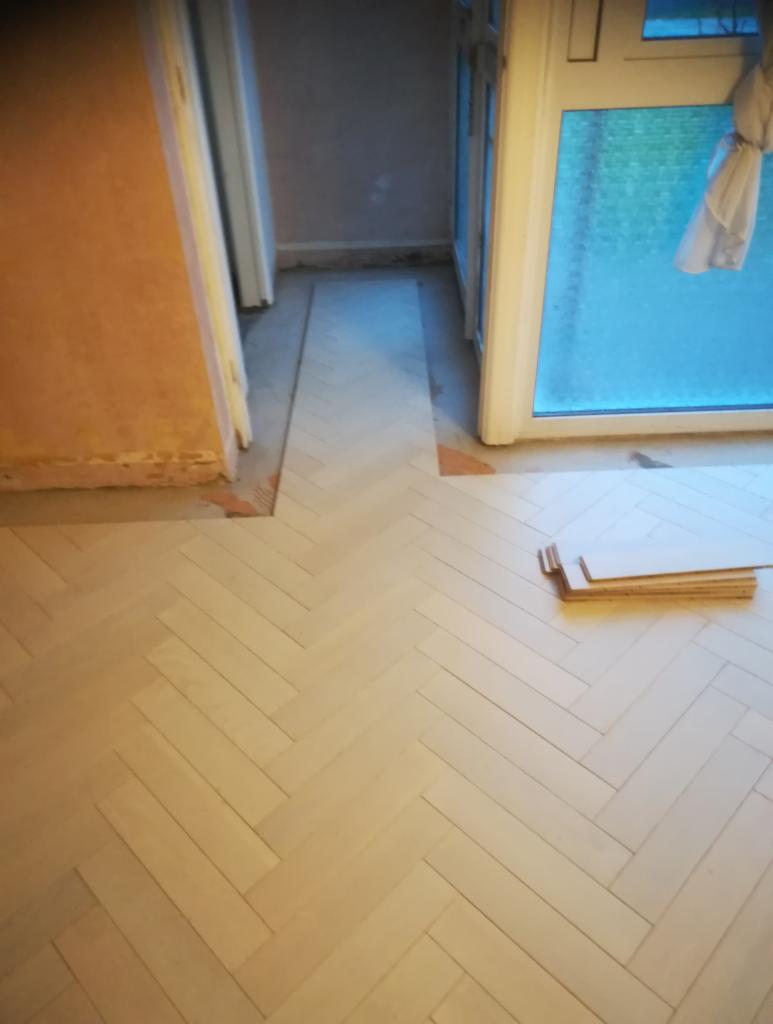 Układanie jasnych paneli podłogowych w jodełkę, widoczne niedokończone fragmenty przy ścianach i paczka paneli obok okna.