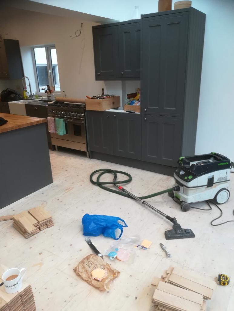 Wnętrze kuchni w trakcie prac wykończeniowych: położona podłoga z paneli, zamontowane szare szafki kuchenne, widoczny odkurzacz przemysłowy, resztki jedzenia i narzędzia na podłodze.