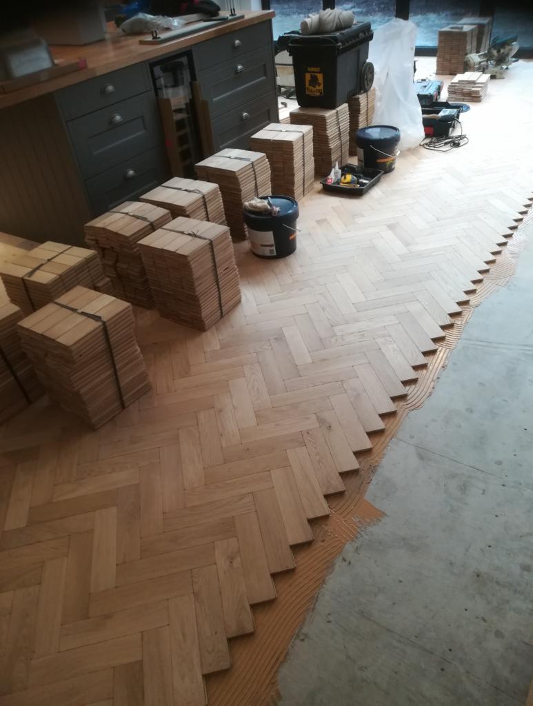 Proces układania parkietu w jodełkę: widoczne paczki desek, klej, oraz częściowo ułożona podłoga w pomieszczeniu z szarą zabudową meblową.