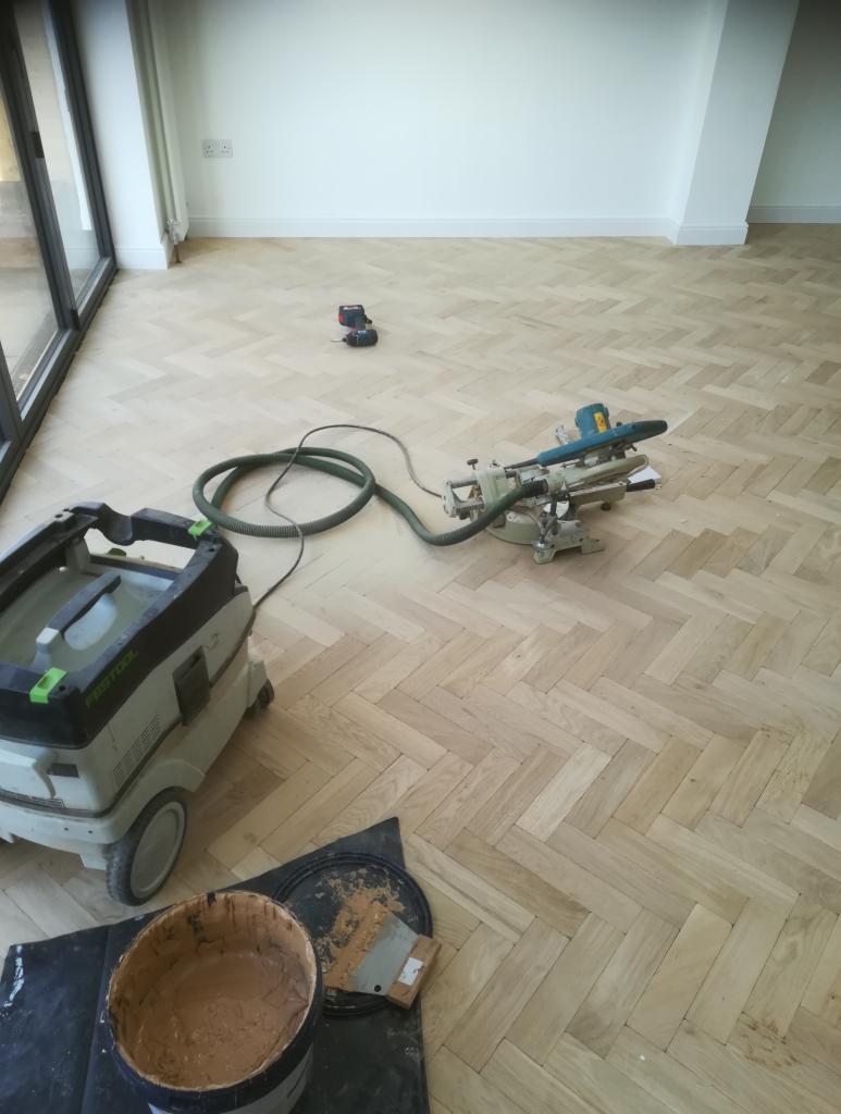 Jasny parkiet ułożony w jodełkę, z widocznym sprzętem budowlanym: odkurzacz przemysłowy Festool, ukośnica i szpachla z wiadrem z masą szpachlową, w tle białe ściany i listwy przypodłogowe.