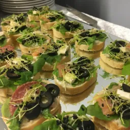 Koreczki na jasnym pieczywie z rukolą, oliwkami, kiełkami i różnymi dodatkami (salami, łosoś, ser brie), ułożone na lustrzanej tacy, widoczne odbicia.
