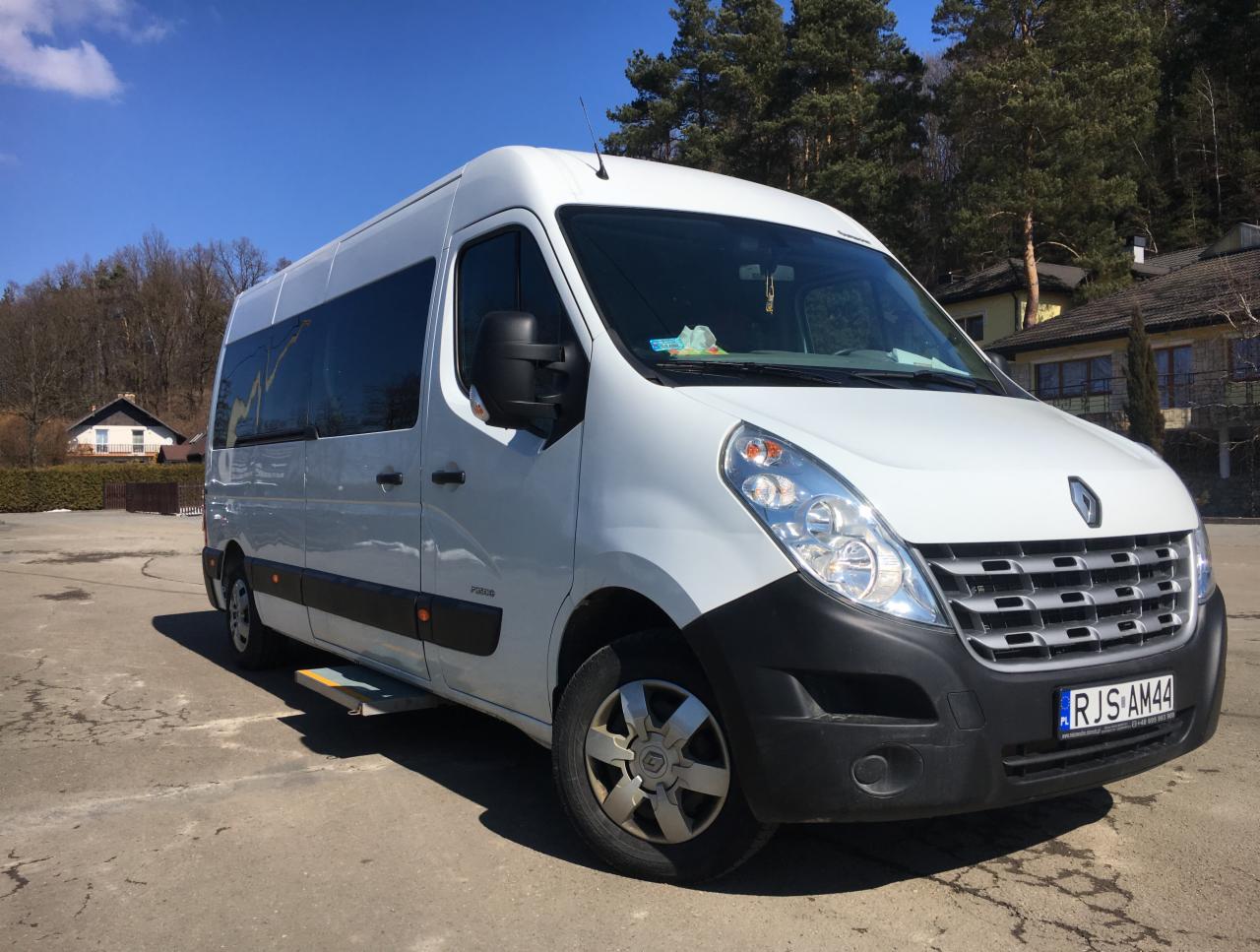 Biały bus Renault Master z wysuwanym stopniem, zaparkowany na betonowym placu w słoneczny dzień, widoczne budynki w tle.