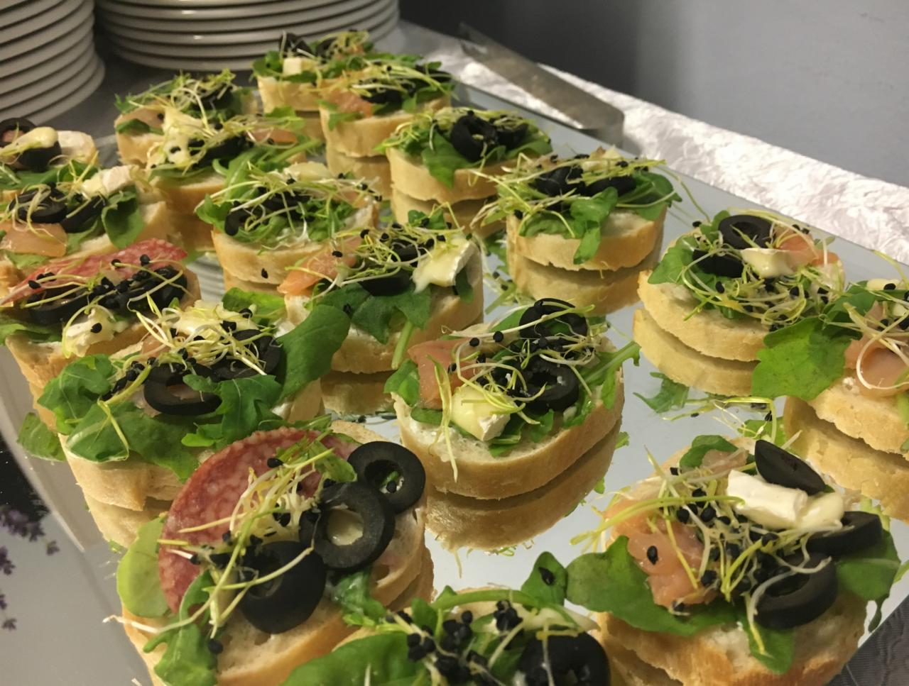 Koreczki na jasnym pieczywie z rukolą, oliwkami, kiełkami i różnymi dodatkami (salami, łosoś, ser brie), ułożone na lustrzanej tacy, widoczne odbicia.