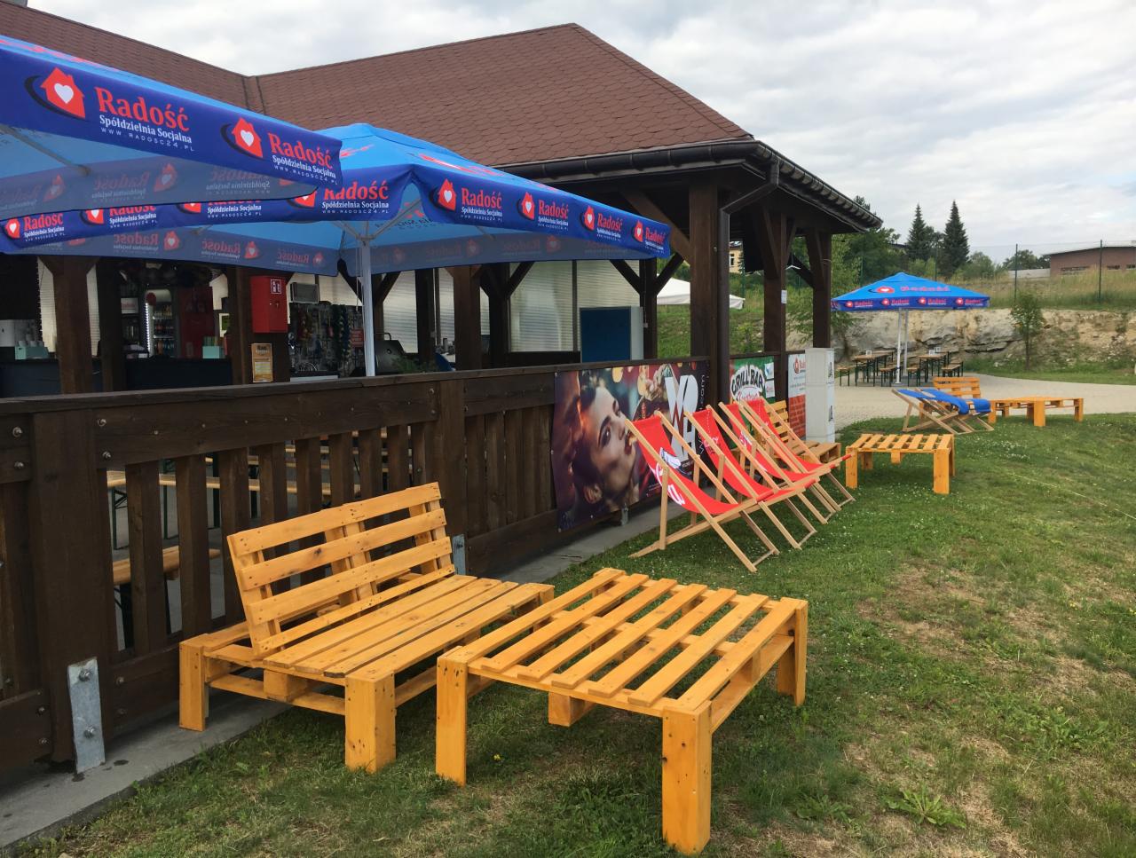 Zewnętrzny bar z parasolami z logo, drewnianymi meblami z palet i leżakami na trawie.