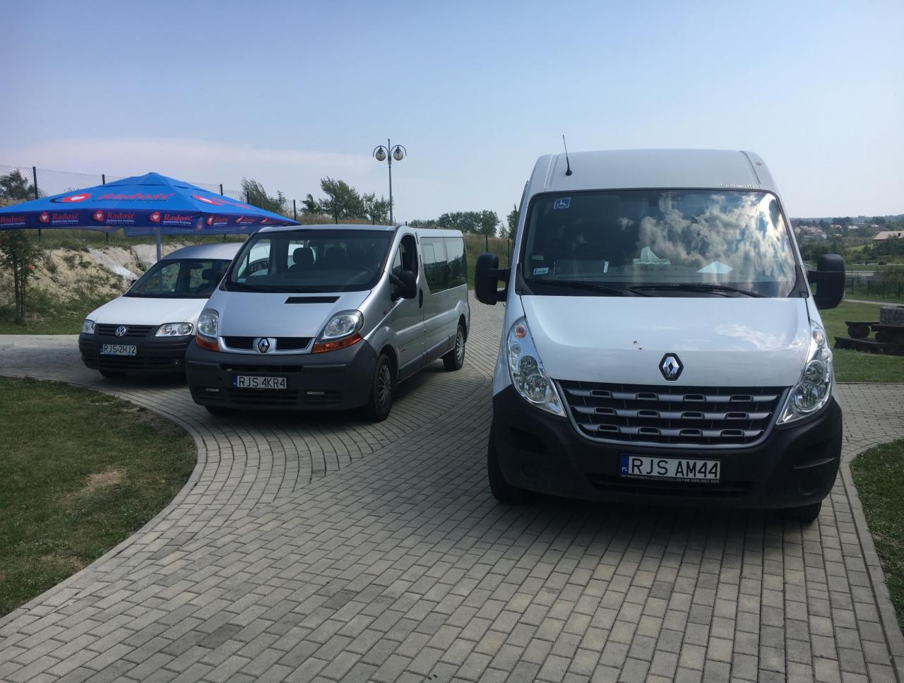 Flota trzech pojazdów: biały Volkswagen, srebrny Renault Trafic i biały Renault Master z symbolem osoby niepełnosprawnej, stojące na brukowanym podjeździe w słoneczny dzień.