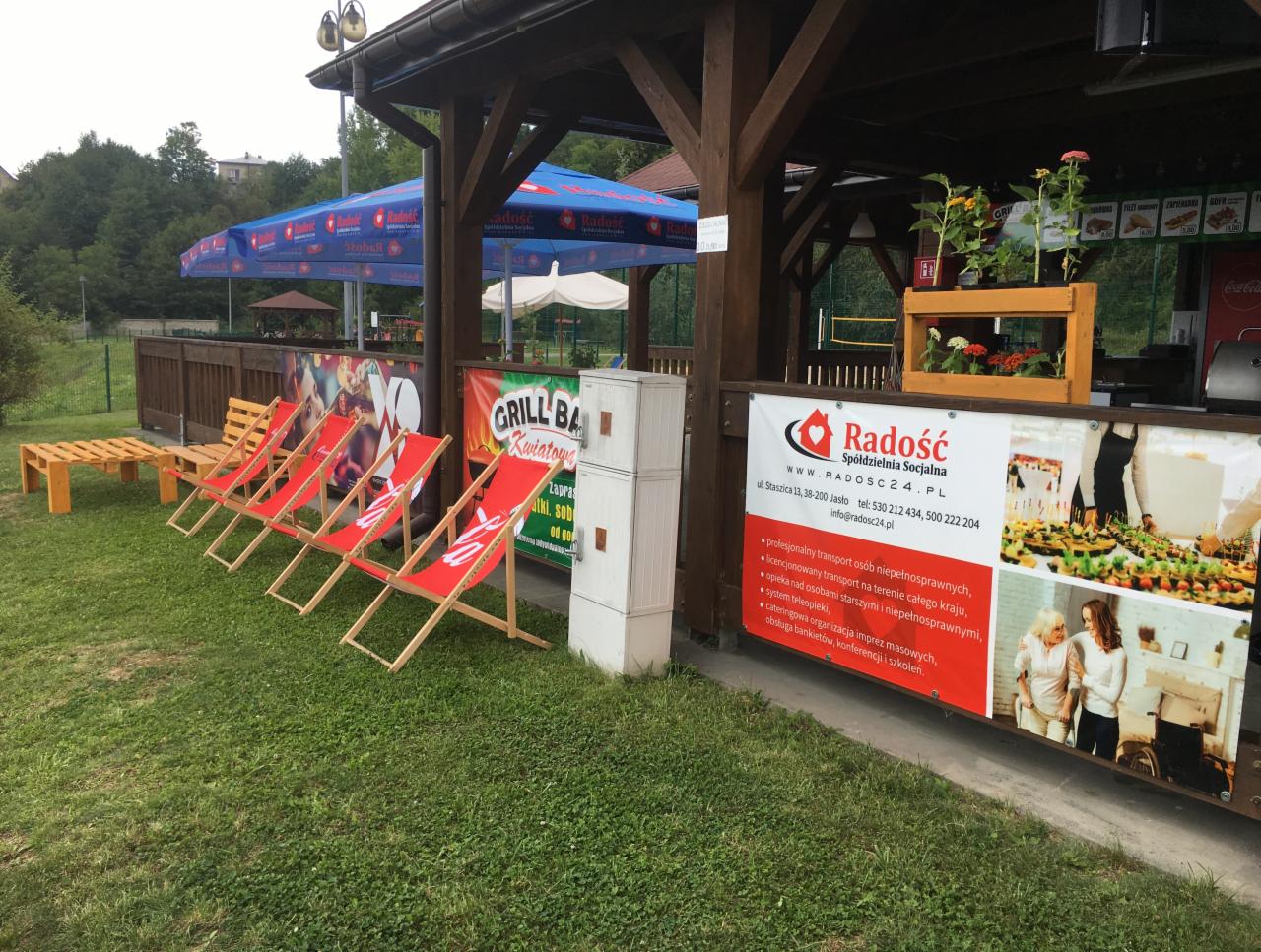 Zewnętrzna altana z czerwonymi leżakami, parasolami i banerem reklamowym Spółdzielni Socjalnej Radość oferującej transport osób niepełnosprawnych, opiekę i catering.