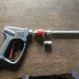 Szara pistoletowa lanca z czerwoną regulacją ciśnienia, elementami złącznymi i dyszą na drewnianym blacie roboczym.