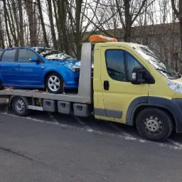 Ż&oacute;łta laweta z niebieskim samochodem osobowym typu kombi na platformie, zaparkowana na parkingu, widoczne drzewa w tle.