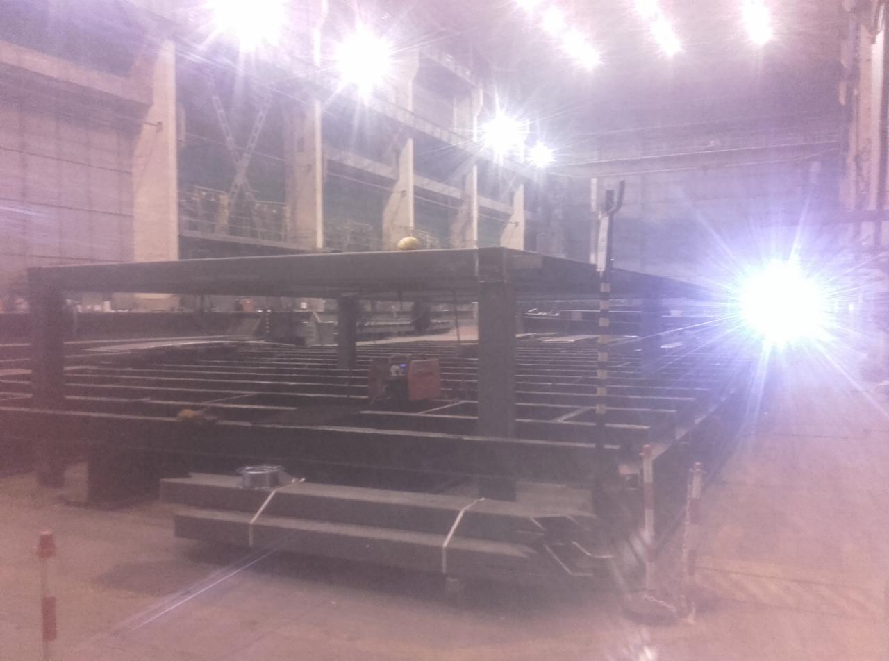 Konstrukcja stalowa w hali produkcyjnej, widoczny szkielet hali, słupy i belki, w tle maszyna spawalnicza, na pierwszym planie stos profili stalowych.