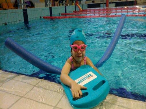 Dziewczynka w różowych okularach pływackich, turkusowym czepku i z niebieską deską do pływania, wsparta na krawędzi basenu z niebieskim makaronem do pływania za plecami.