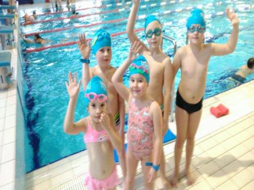 Grupa dzieci w czepkach i strojach kąpielowych macha rękami na tle basenu, w tle widoczni inni pływacy.