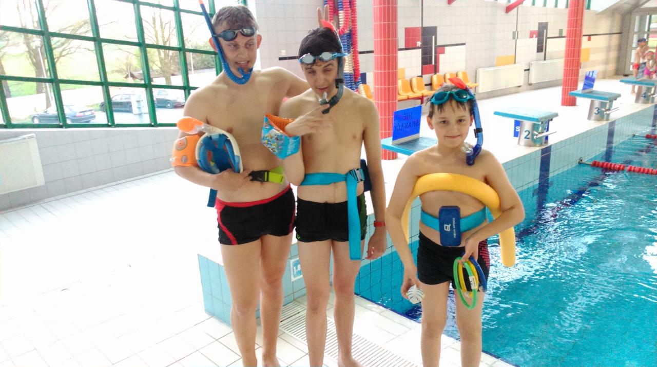 Trzech chłopców w strojach kąpielowych z rurkami i okularami, stojących na brzegu basenu, z różnymi akcesoriami do pływania, w tym rękawkami i pasami wypornościowymi.