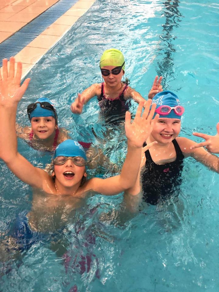 Grupa uśmiechniętych dzieci w czepkach i okularach pływackich, machających rękami w basenie w Jastrzębiu-Zdroju, podczas zajęć nauki pływania.