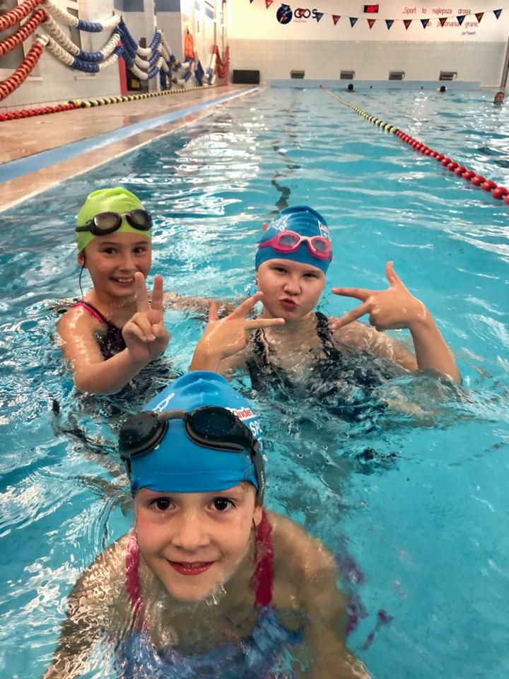 Trzy uśmiechnięte dziewczynki w czepkach i okularach pływackich pozują w basenie, jedna z nich pokazuje znak 'V', w tle widoczne liny oddzielające tory i dekoracje na ścianie.