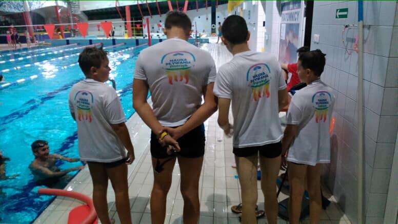 Grupa czterech chłopców w białych koszulkach z logo na plecach, stojących na brzegu basenu krytego. W tle widoczni inni pływający.