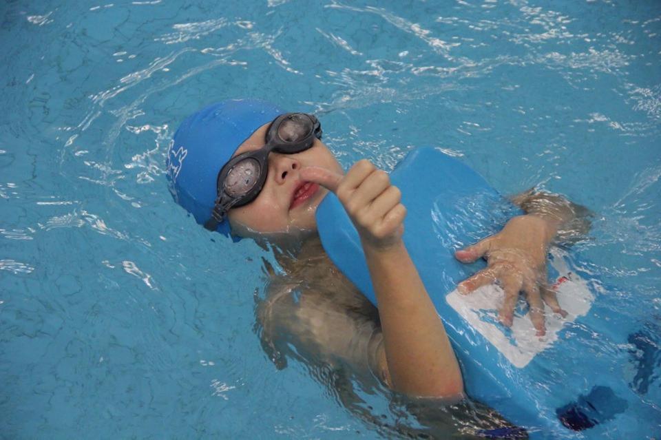 Chłopiec w czepku i okularach pływackich unosi się na wodzie, trzymając niebieską deskę do pływania, z palcem wskazującym przy ustach.