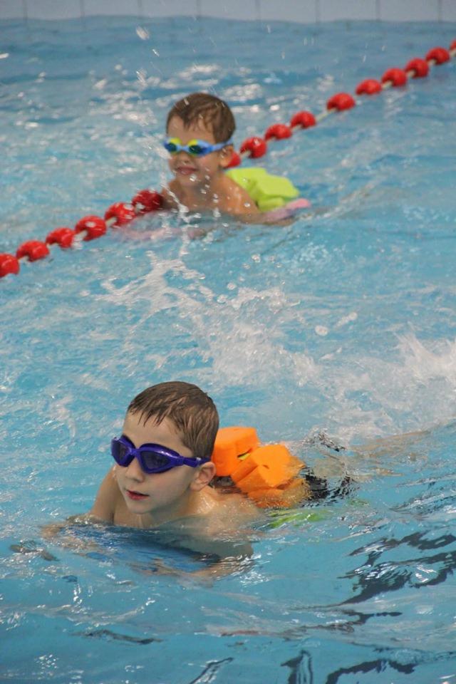 Dwaj chłopcy w okularach pływackich i z piankowymi wyporami na plecach unoszą się na wodzie w basenie, oddzieleni czerwonymi bojami.