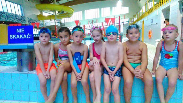 Grupa dzieci w strojach kąpielowych i goglach siedzi na krawędzi basenu, w tle widoczny znak zakazu skakania do wody oraz zjeżdżalnia wodna.