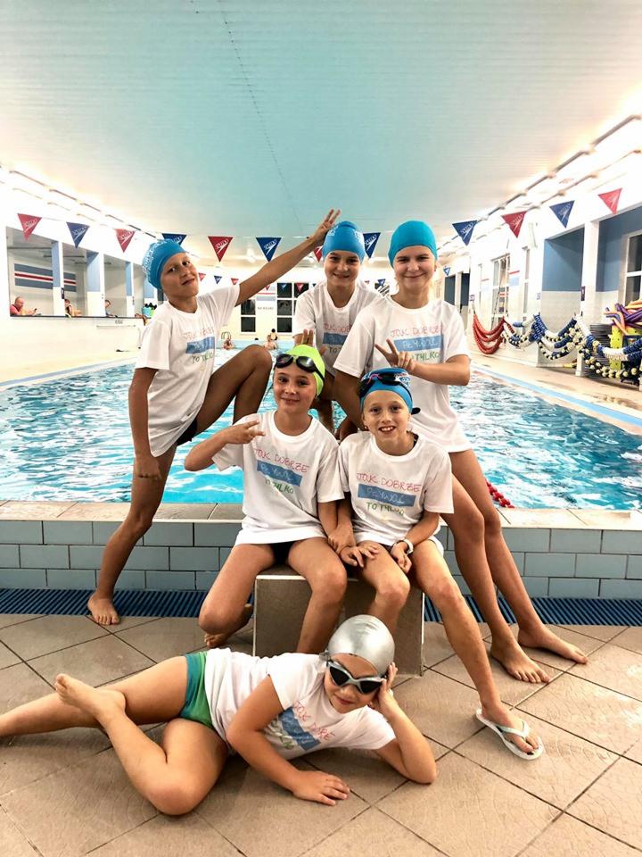 Grupa uśmiechniętych dzieci w czepkach i koszulkach z nadrukiem pozuje na tle basenu, jedno dziecko leży na podłodze w okularach pływackich i czepku, pozostałe stoją lub siedzą na białym postumencie.