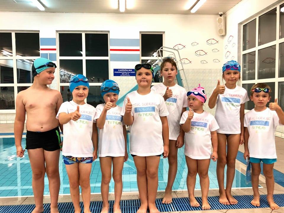 Grupa dzieci w strojach kąpielowych i koszulkach z napisem 'Jak dobrze pływać to tylko' stoi na brzegu basenu, wszyscy pokazują kciuki w górę.