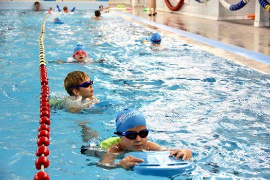 Grupa dzieci w czepkach i okularkach pływackich w basenie, używających desek do pływania, widoczna lina oddzielająca tory.