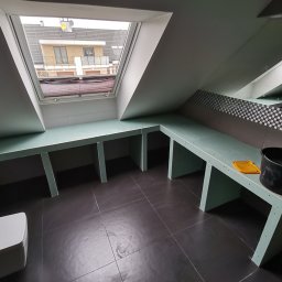 Wróbel Build Group - Montaż płyt karton gips w łazience na poddaszu we Wrocławiu, widoczne stelaże pod zabudowę umywalki i toalety, czarne płytki na podłodze, okno dachowe.