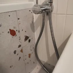 Bateria prysznicowa marki Tres zamontowana na ścianie z płytkami w stylu lastryko, obok fragmentu białej wanny.