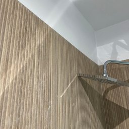 Wróbel Build Group - Wykończona ściana w kabinie prysznicowej z panelami imitującymi drewno, chromowana deszczownica zamontowana pod sufitem.