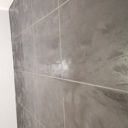 Wróbel Build Group - Ściana wykończona szarymi płytkami imitującymi beton, z widocznymi fugami i fragmentem białej ściany oraz podłogi z paneli.