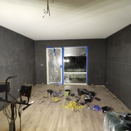 Wróbel Build Group - Pokój po malowaniu ścian na ciemny kolor, z widocznymi śladami taśmy malarskiej na podłodze i zabezpieczonymi folią oknami, oświetlony lampami budowlanymi.