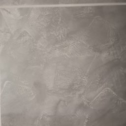 Wróbel Build Group - Szara ściana z dekoracyjną fakturą gładzi, obramowana taśmą malarską, ukazująca nieregularne wzory i subtelne cieniowanie.