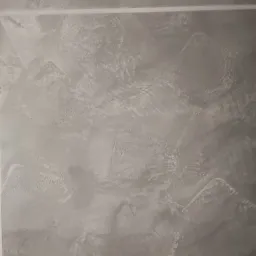 Szara ściana z dekoracyjną fakturą gładzi, obramowana taśmą malarską, ukazująca nieregularne wzory i subtelne cieniowanie.