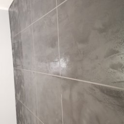 Wróbel Build Group - Ściana wyłożona szarymi, kwadratowymi płytkami z jasną fugą, widoczne niedoskonałości i nieregularna faktura na powierzchni płytek, przy krawędzi ściany widoczny fragment podłogi z paneli.