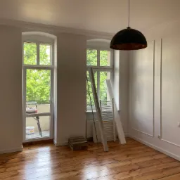 Pomalowane na biało wnętrze pokoju z drewnianą podłogą, widoczne okna z zielenią na zewnątrz, drabina oparta o ścianę oraz wisząca lampa z czarnym kloszem.