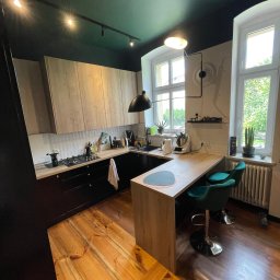 Wróbel Build Group - Nowoczesna kuchnia z drewnianymi szafkami, czarnymi elementami i zielonymi krzesłami barowymi, oświetlona punktowymi lampami na ciemnozielonym suficie. W tle dwa okna z widokiem na zieleń.