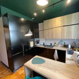 Wróbel Build Group - Nowoczesna kuchnia z ciemnozielonym sufitem, czarnymi szafkami i drewnianymi blatami, oświetlona reflektorami. Widoczny fragment wyspy kuchennej z zielonym hokerym.