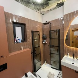 Wróbel Build Group - Nowoczesna łazienka po remoncie z brązowymi płytkami, kabiną prysznicową typu walk-in z czarnymi ramami, umywalką z szafką w kolorze terakoty i bidetem; sufit zdobi tapeta z motywem tropikalnych...