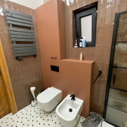 Wróbel Build Group - Nowoczesna łazienka z brązowymi, pionowo ułożonymi płytkami, białym sedesem i bidetem z czarną armaturą, grzejnikiem drabinkowym oraz szafką w kolorze terakoty.