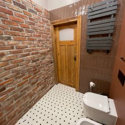 Wróbel Build Group - Wykończona łazienka z ceglaną ścianą, białą mozaikową podłogą w czarne kropki, drewnianymi drzwiami i grzejnikiem drabinkowym w kolorze szarym.