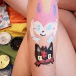 Ręka z pomalowanymi farbami postaciami z bajek, widoczne farby do malowania twarzy i tęczowe tło.