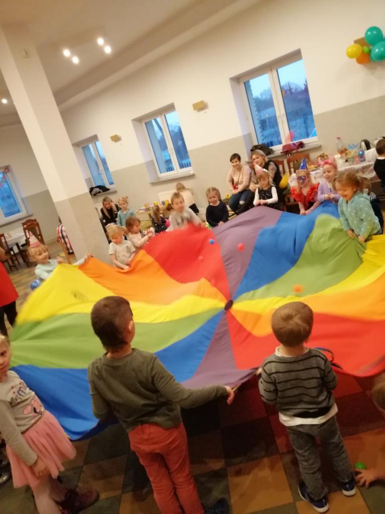 Grupa dzieci bawi się kolorową chustą animacyjną w dużej sali z oknami, pod nadzorem dorosłych.