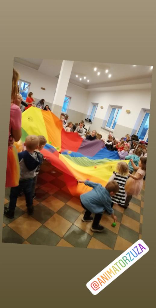 Grupa dzieci bawi się kolorową chustą animacyjną w przestronnym pomieszczeniu z kafelkową podłogą i oknami.